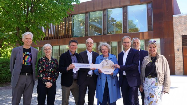 Acht Menschen stehen vor der neuen Bibliothek. In der Mitte wird von drei Personen ein Übergabeschlüssel gehalten.