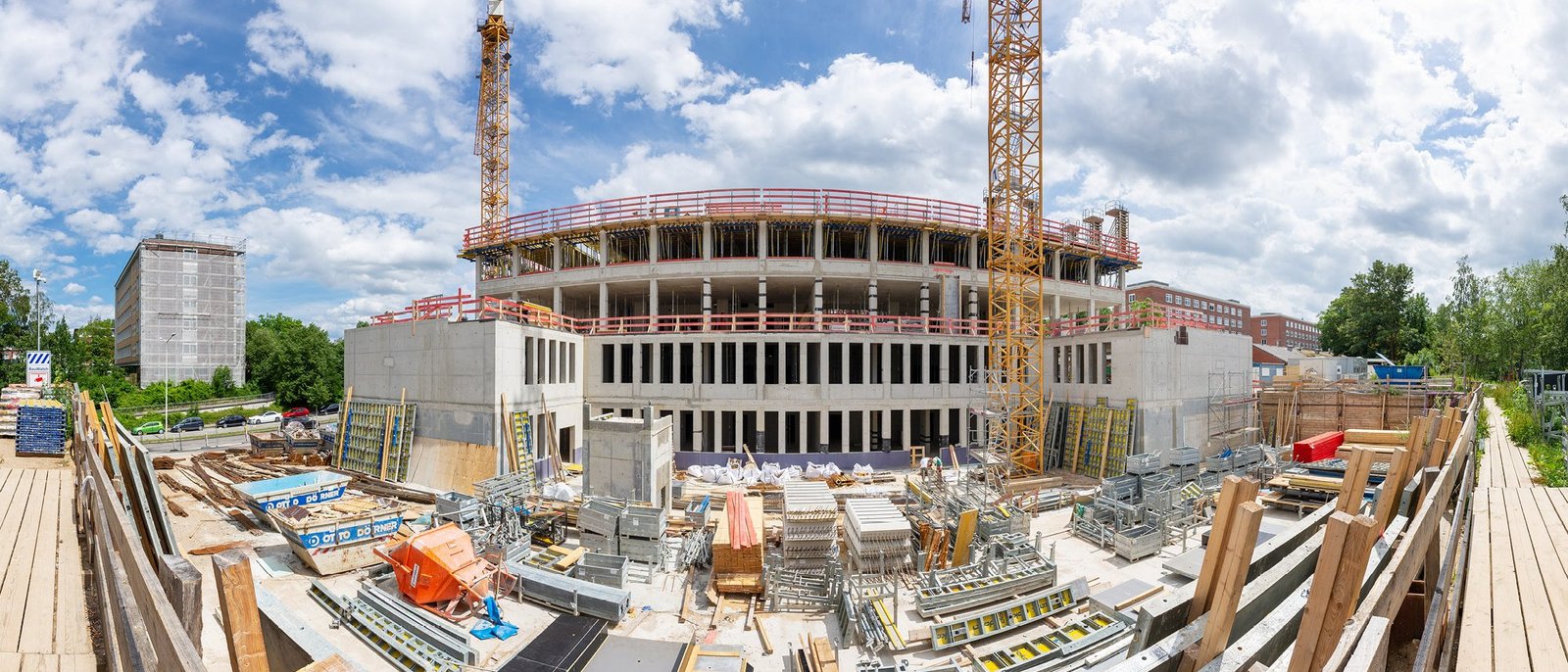 Panoramabild Baustelle Geowissenschaftliches Institut. Blick ins Foyer. Vier unfertige Geschosse mit Fensteraussparungen sind zu sehen. Auf dem Betonboden liegen viele Stahlteile. Links und rechts stehen zwei Kräne, die halb zu sehen sind. Oben ist blauer Himmel.