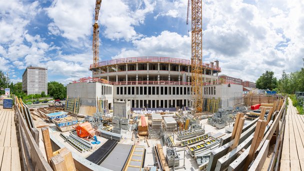 Panoramabild Baustelle Geowissenschaftliches Institut. Blick ins Foyer. Vier unfertige Geschosse mit Fensteraussparungen sind zu sehen. Auf dem Betonboden liegen viele Stahlteile. Links und rechts stehen zwei Kräne, die halb zu sehen sind. Oben ist blauer Himmel.