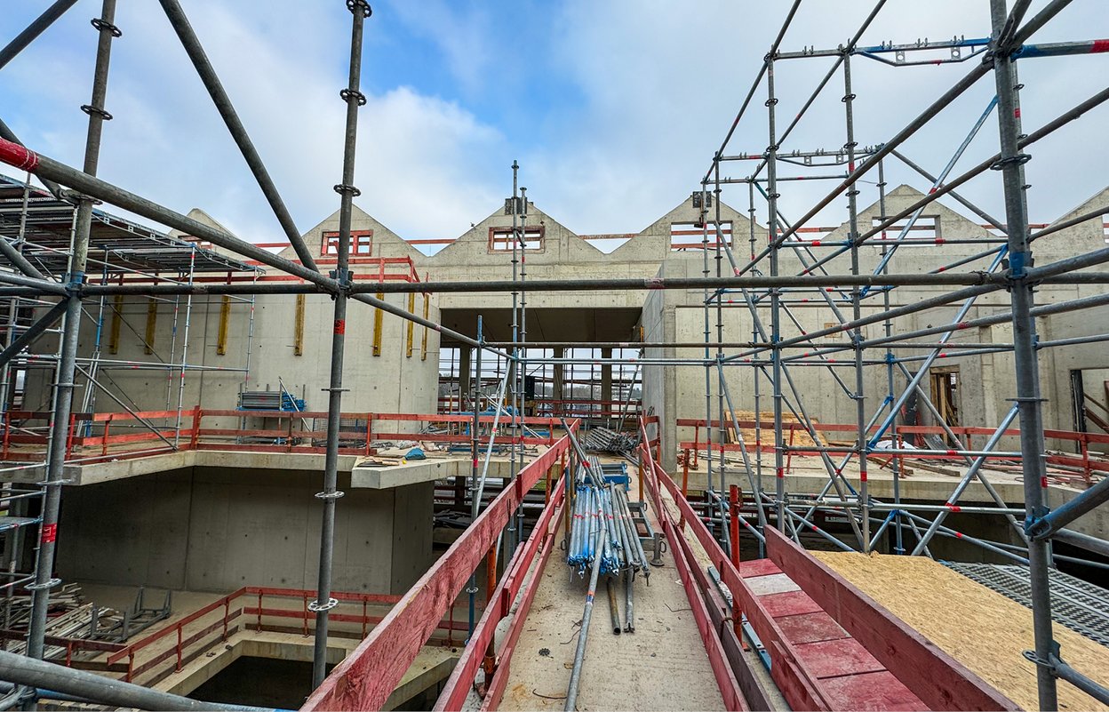 Baustelle eines modernen Betongebäudes mit Gerüsten und provisorischer Rampe im Rohbauzustand unter leicht bewölktem Himmel.