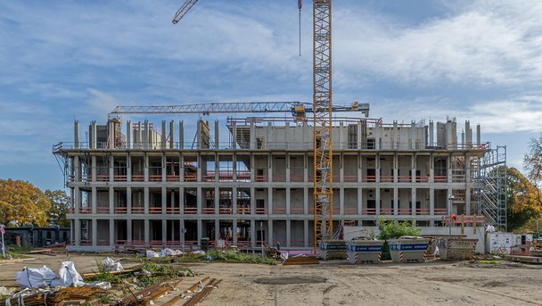 Mehrstöckiger Rohbau eines Betongebäudes mit Baukränen und Gerüsten, umgeben von Baumaterialien und Containern auf einer offenen Baustelle.