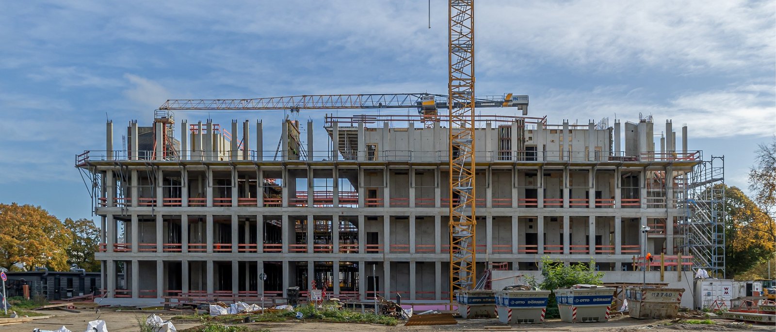 Mehrstöckiger Rohbau eines Betongebäudes mit Baukränen und Gerüsten, umgeben von Baumaterialien und Containern auf einer offenen Baustelle.