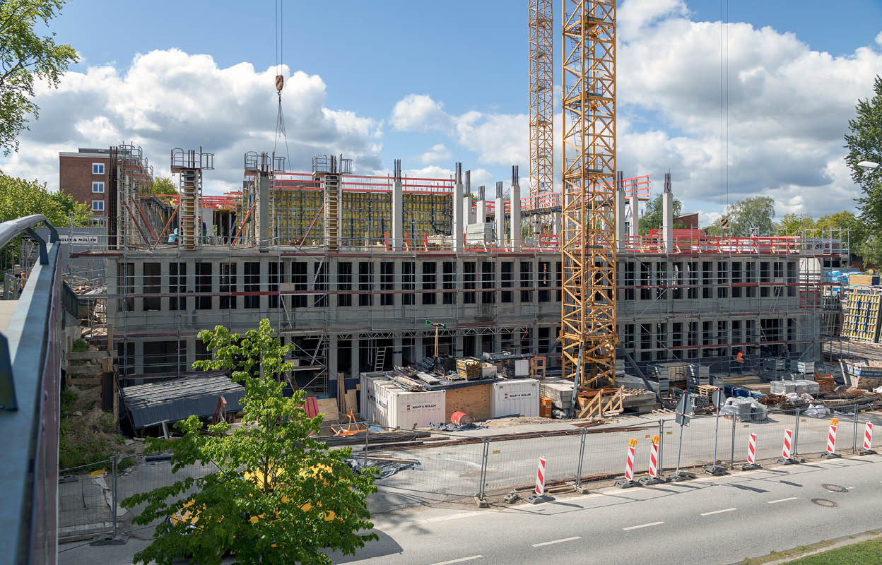 Baustelle Geowissenschaftliches Institut von außen betrachtet. Links ist eine Fußgängerbrücke zu sehen. Rechts ein Kran. Mittig sind das erste und zweite Geschoß zu sehen. Es wird gerade an den Stahlbetonstützen für das dritte Geschoß gearbeitet. Oben ist blauer Himmel zu sehen. 