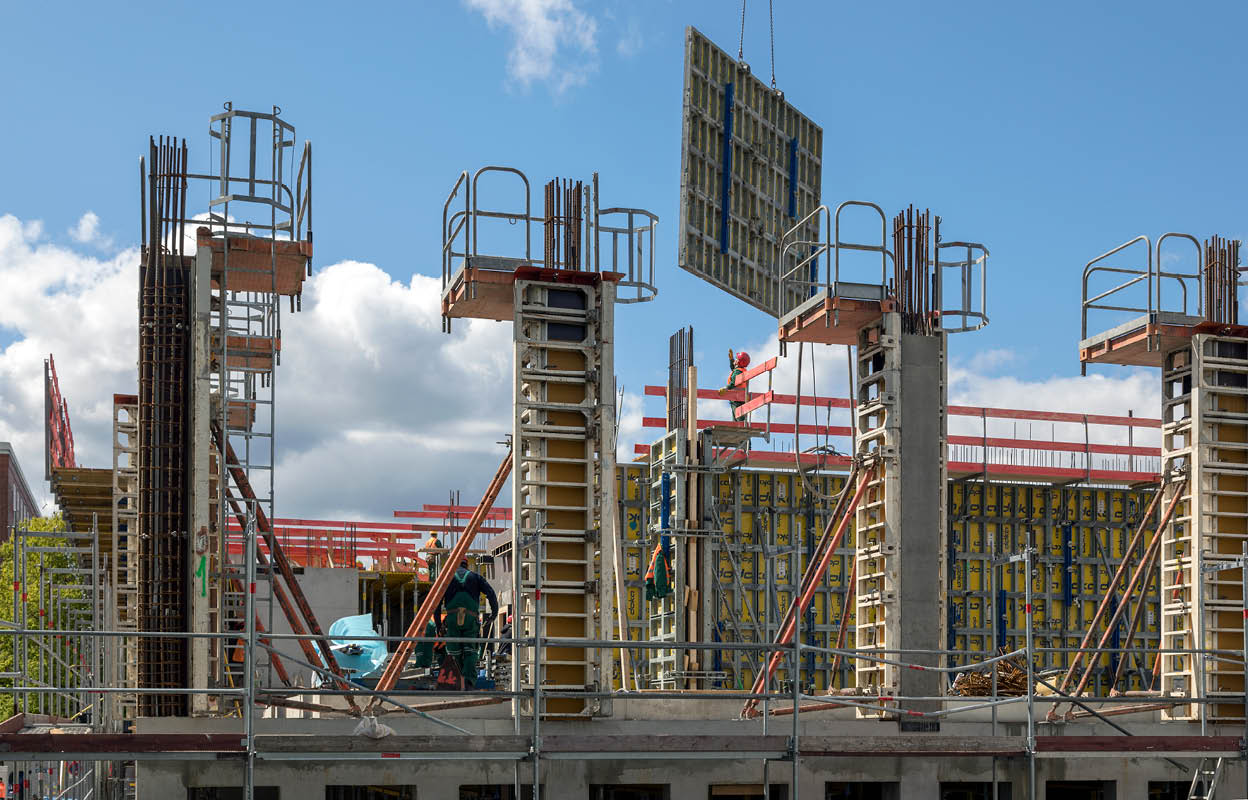 Baustelle Geowissenschaftliches Institut. Es sind vier Stahlbetonstützen zu sehen. An einem Kran hängt eine vorgefertigte Zwischenwand. Oben ist blauer Himmel zu sehen. 