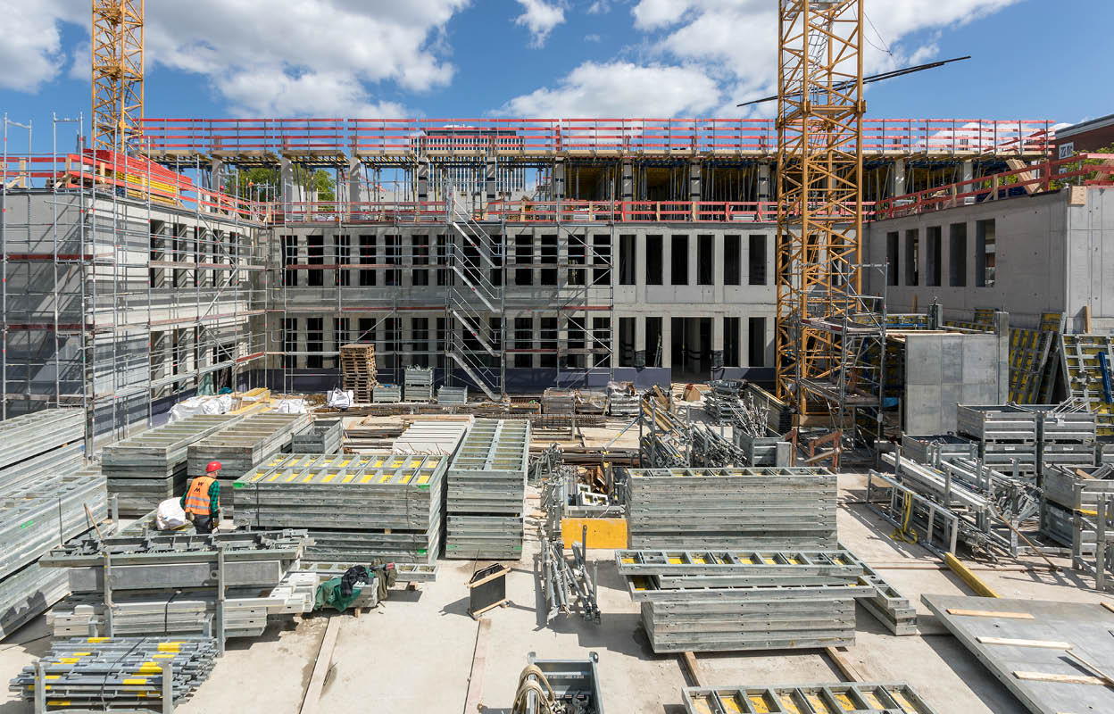 Baustelle Geowissenschaftliches Institut. Blick ins Foyer. Zwei unfertige Geschosse mit Fensterlücken sind zu sehen. Auf dem Betonboden liegen viele Stahlteile. Links und rechts stehen zwei Kräne, die halb zu sehen sind. Oben ist blauer Himmel.